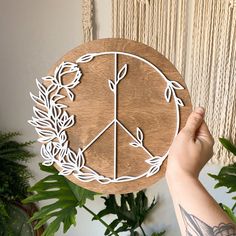 a person holding up a wooden peace sign in front of a plant and wall hanging