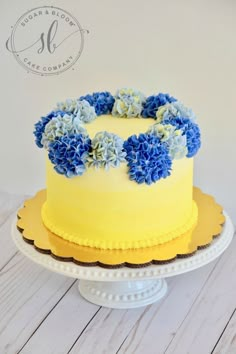 a yellow cake with blue flowers on top sitting on a white plate next to a wooden table