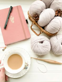 a person holding a cup of coffee next to yarn and a notebook on a table