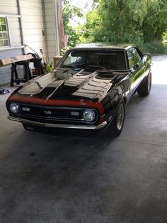 a black and red muscle car parked in a garage next to a building with trees