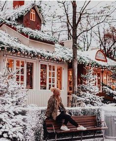 a person sitting on a bench in front of a house with snow covered trees and bushes