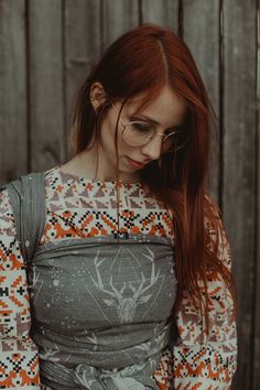 a woman with red hair wearing glasses and a patterned top standing in front of a wooden fence