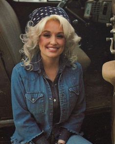 a woman sitting on the ground next to a car wearing a jean jacket and polka dot headband