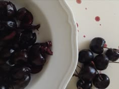 some black cherries are in a white bowl on a table next to another plate