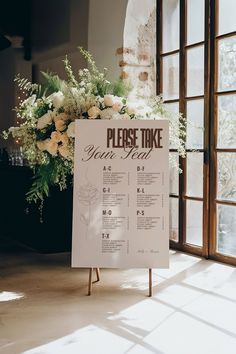 a sign that says please take your seat next to a bouquet of flowers and greenery