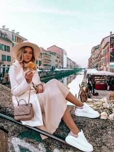 a woman in a white dress and hat sitting on the edge of a bridge eating food