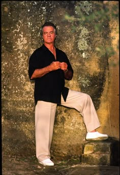 a man in white pants and black shirt leaning against a stone wall