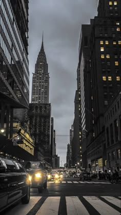 cars driving down a city street at night with tall buildings in the background and lights on