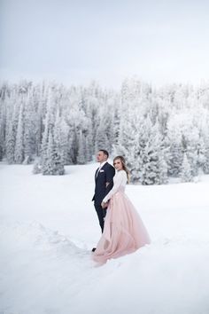 a man and woman standing in the snow with trees behind them, holding each other