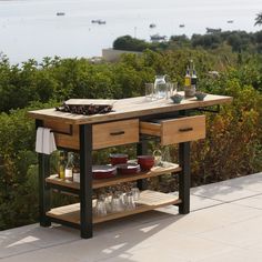 an outdoor table with drinks and glasses on it near some bushes, overlooking the water