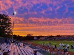 a group of people standing on top of a baseball field under a colorful sky at sunset