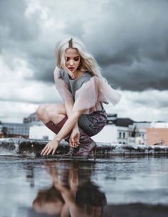 a woman crouching down on the ground in front of a body of water with buildings behind her