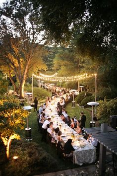 a long table is set up in the middle of a garden with lights strung over it