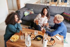 three people sitting at a table eating pizza and drinking sodas while another person sits on the couch