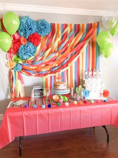 a table topped with cake and balloons next to a wall covered in colorful streamers