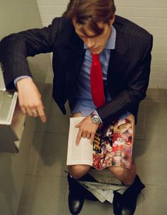 a man in a suit and tie sitting on a toilet reading a book while wearing shorts