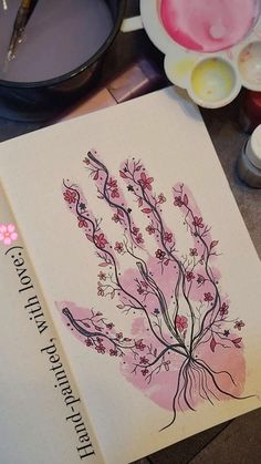 an open book with pink flowers on it next to paint and other items in the background