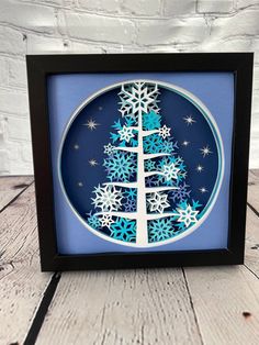 a paper cut christmas tree with snowflakes on it in a black frame against a white brick wall