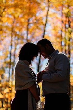 a man and woman standing next to each other in front of trees with yellow leaves