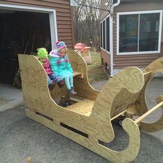 two children are sitting in a wooden sleigh on the driveway next to their garage