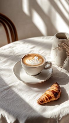 a cappuccino and croissant sitting on a white tablecloth next to a cup of coffee