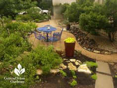 a table and chairs are in the middle of a garden with rocks, plants, and gravel