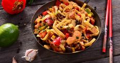 a bowl filled with pasta and vegetables on top of a wooden table next to chopsticks