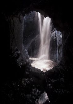 a waterfall is coming out of a cave into the dark water with light streaming from it