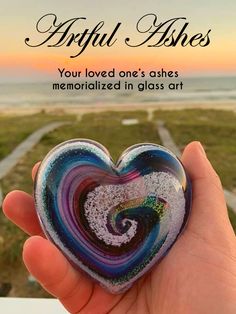 a hand holding a glass heart in front of the ocean with text that reads, actual ashes your loved one's otherwise memorialized in glass art