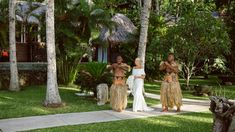 three women dressed in hula skirts are standing on the grass near trees and palm trees