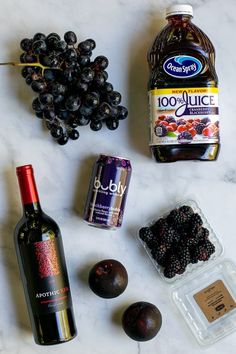 various fruits and condiments are laid out on a marble counter top, including plums, blackberries, olives, an orange juice