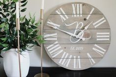 a wooden clock with roman numerals on it next to a potted plant