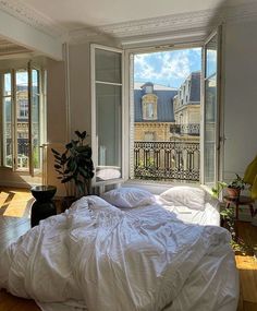 an unmade bed with white sheets and pillows in front of two large windows overlooking the city