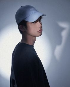 a young man wearing a blue hat and black t - shirt, standing in front of a white wall