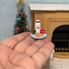 a hand is holding a small plate with cookies and milk on it in front of a miniature christmas tree