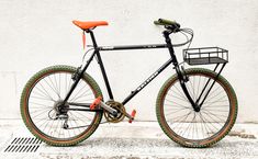 a black bike with orange rims parked next to a white wall and drain grate