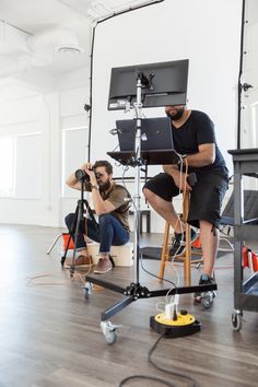 two men sitting on stools with laptops in front of them and one man standing behind the camera