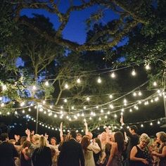 a group of people standing around each other at a party with lights strung from the trees