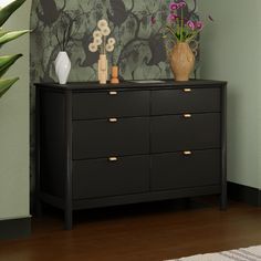 a black dresser with flowers and vases on it in front of a green wall