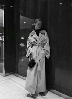 a woman standing in front of a wall wearing a trench coat and holding a purse