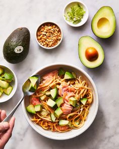 someone holding a spoon over a bowl of noodles with avocado and other ingredients