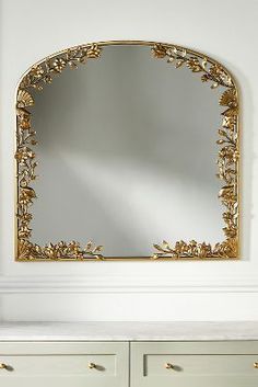 an ornate gold framed mirror on a white wall above a dresser with drawers and vase