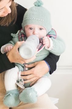 a woman is holding a baby and drinking from a bottle while wearing a knitted hat