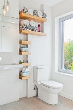 a white toilet sitting next to a window in a bathroom under a shelf filled with dishes