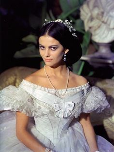 a woman wearing a white dress and tiara sitting in front of a flower arrangement