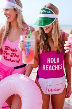 two women in bathing suits on the beach with drinks and floaters, one holding a drink