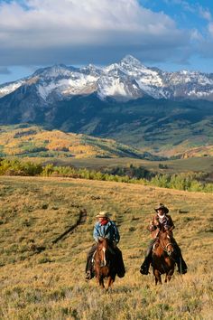 two people are riding horses in an open field with mountains in the backgroud