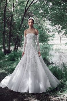 a woman in a white wedding dress standing on the ground near trees and grass, with her arms behind her back