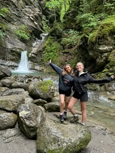 two young women standing on rocks in front of a waterfall with their arms spread out