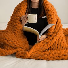 a woman is reading a book and holding a coffee cup while wrapped in a blanket